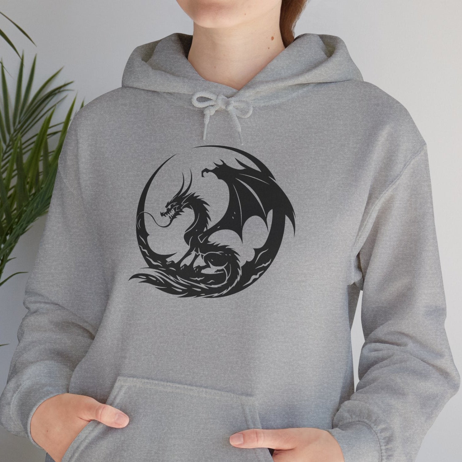 Person wearing a gray hoodie with a black dragon design in a room with a plant.